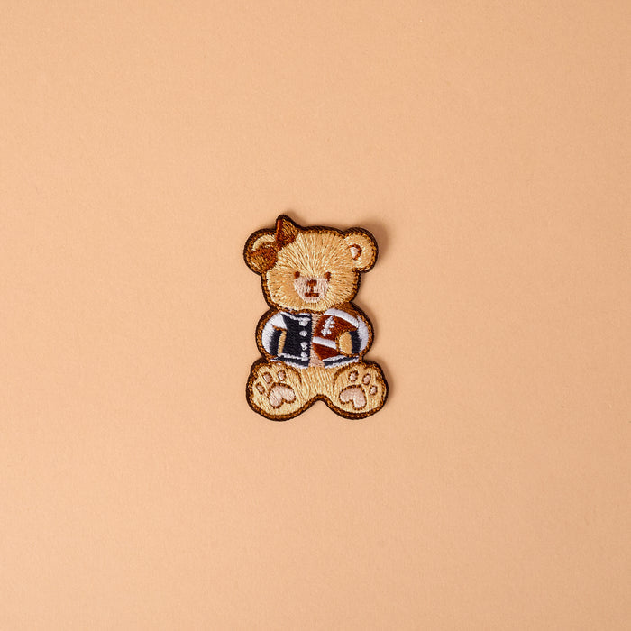 The Gameday Touchdown Teddy Mix & Match Iron-On Patch by The Darling Effect features an embroidered brown teddy bear dressed in a blue jacket and holding a small brown item, set against a plain beige background—ideal for anyone who loves customizable patches or wants to add unique iron-on designs to their collection.
