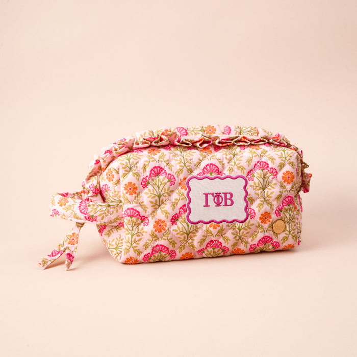 The Darling Effect’s Gamma Phi Beta Essentials Quilted Cosmetic Pouch features pink, orange, and green floral patterns, a decorative “ΓΦΒ” label in the center, and a tied fabric handle—ideal for organizing your sorority essentials.