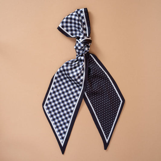 The Darling Effect’s Gingham Black Twirl & Tie Scarf Accessory is a chic black-and-white satin scarf featuring a gingham pattern on one side and playful polka dots on the other, shown tied in a loose knot against a beige background—an elegant and versatile accessory that effortlessly elevates any wardrobe.