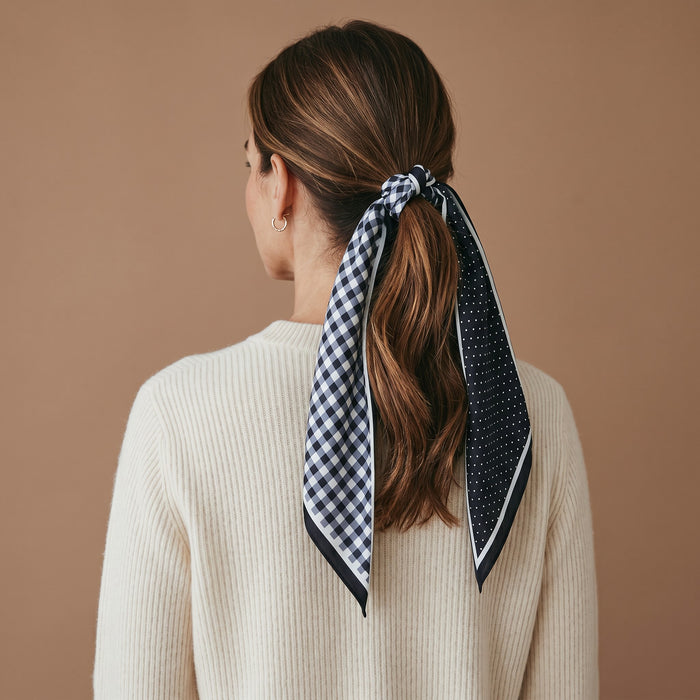 A woman with brown hair styled in a ponytail, tied with The Darling Effect’s Gingham Black Twirl & Tie Scarf Accessory—a black and white checkered satin scarf featuring polka dot details—wears a cream sweater and is shown from behind against a plain brown background.