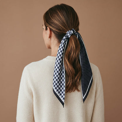 A woman with brown hair styled in a ponytail, tied with The Darling Effect’s Gingham Black Twirl & Tie Scarf Accessory—a black and white checkered satin scarf featuring polka dot details—wears a cream sweater and is shown from behind against a plain brown background.