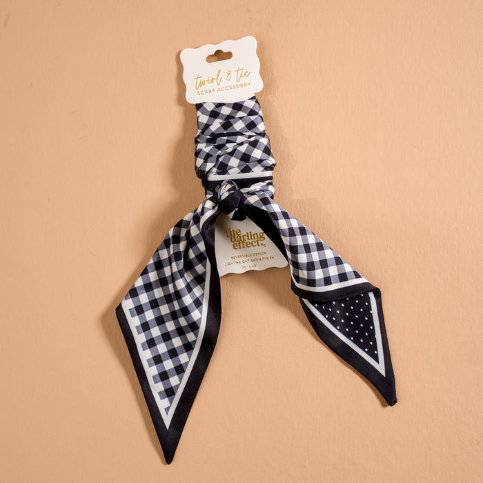 The Darling Effect’s Gingham Black Twirl & Tie Scarf Accessory is shown on a beige background, featuring a classic black and white check pattern and offering versatile styling options for any outfit.