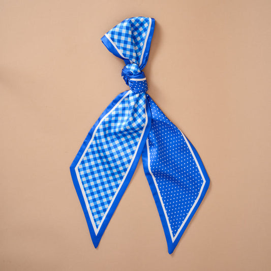 The Darling Effect's Gingham Blue Twirl & Tie Scarf Accessory features blue satin with one side in a gingham pattern and the other in polka dots, both framed by a crisp white border. Shown tied in a loose knot on a beige background, this elegant scarf offers versatile styling options.