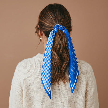 A person with wavy brown hair wears The Darling Effect's Gingham Blue Twirl & Tie Scarf Accessory, styled in their ponytail. Paired with a cream sweater and photographed from behind against a tan background, the image highlights creative ways to accessorize with this unique blue and white patterned scarf.