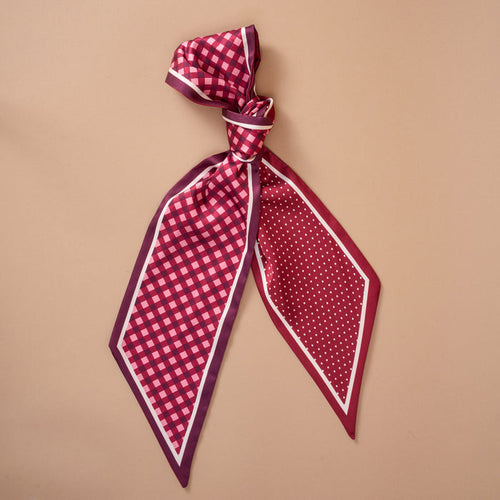 The Gingham Burgundy Twirl & Tie Scarf Accessory from The Darling Effect features one end with a pink checkered pattern and the other adorned with white polka dots on a red background, shown tied in a knot on a beige surface—perfect for experimenting with various styling options.