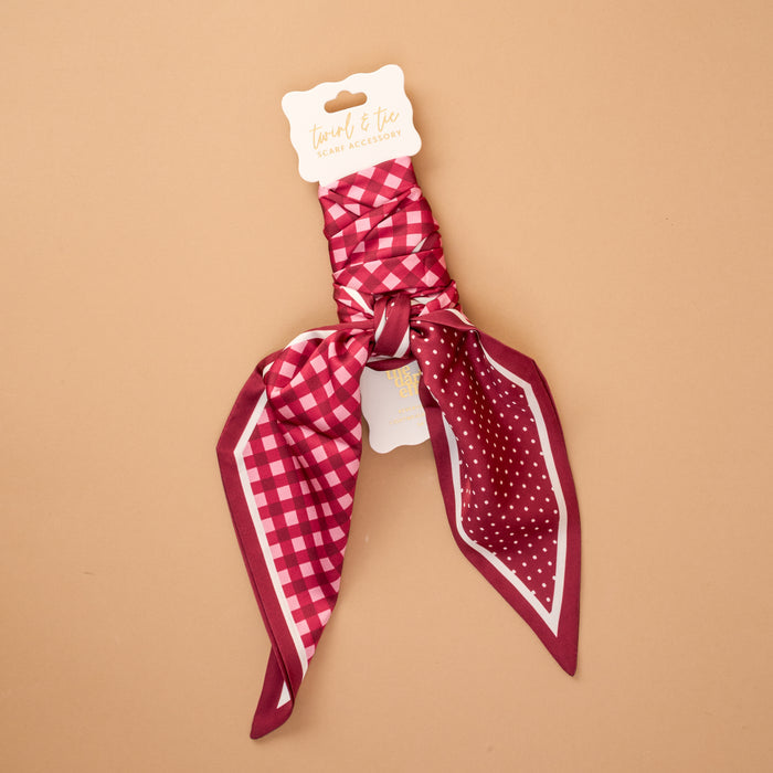 A Gingham Burgundy Twirl & Tie Scarf Accessory by The Darling Effect featuring a maroon, pink, and white satin design with checks and polka dots is neatly tied and attached to a product tag labeled "twist & tie great accessory," displayed against a tan background. This versatile scarf offers endless styling options to complement any outfit.