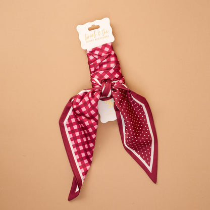 A Gingham Burgundy Twirl & Tie Scarf Accessory by The Darling Effect featuring a maroon, pink, and white satin design with checks and polka dots is neatly tied and attached to a product tag labeled "twist & tie great accessory," displayed against a tan background. This versatile scarf offers endless styling options to complement any outfit.