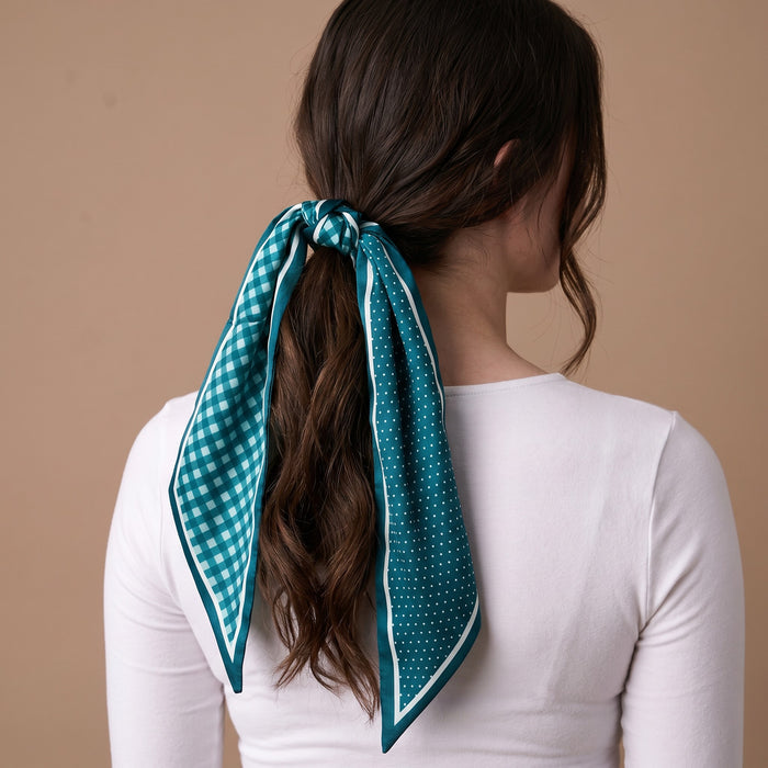 A woman with wavy brown hair, shown from behind, wears The Darling Effect’s Gingham Green Twirl & Tie Scarf Accessory tied around her ponytail. Demonstrating chic styling options, she pairs it with a white top against a plain beige background.