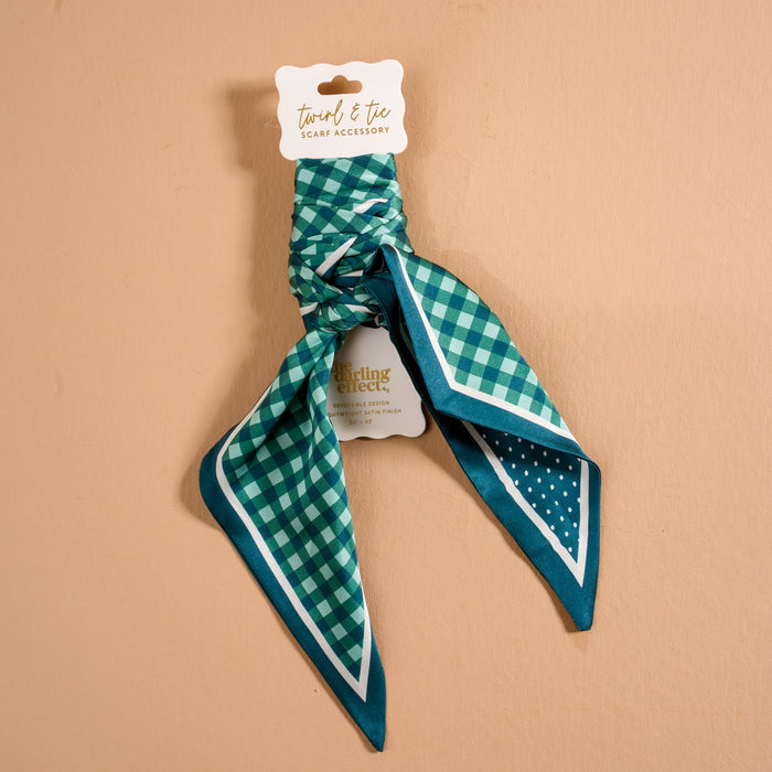 A Gingham Green Twirl & Tie Scarf Accessory by The Darling Effect, featuring teal, navy, and white checkered patterns with solid and polka dot borders, is tied around a cardboard display labeled "twirl & tie scarf accessory" and set against a beige background to showcase versatile styling options.