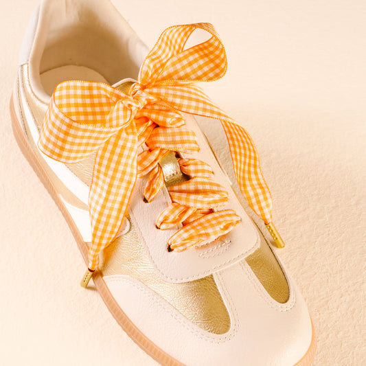 The Darling Effect's Gingham Orange Step It Up Shoelaces, tied in a large bow on a white and gold sneaker, are showcased against a light beige background.