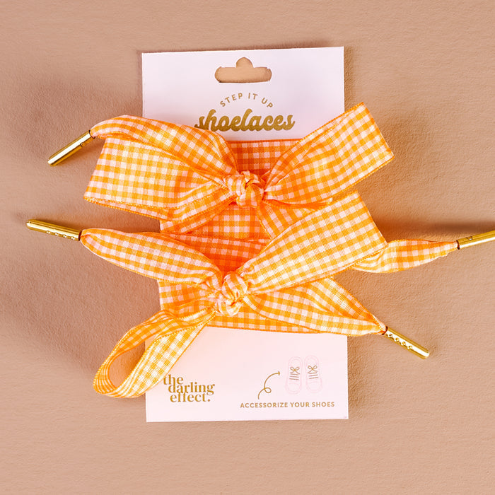 Two orange and white gingham Gingham Orange Step It Up Shoelaces by The Darling Effect, finished with gold tips, are tied in bows and displayed on a branded card. Durable laces to accessorize your shoes with fresh, stylish flair.