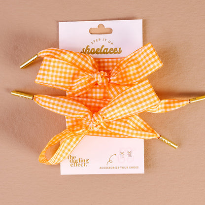 Two orange and white gingham Gingham Orange Step It Up Shoelaces by The Darling Effect, finished with gold tips, are tied in bows and displayed on a branded card. Durable laces to accessorize your shoes with fresh, stylish flair.