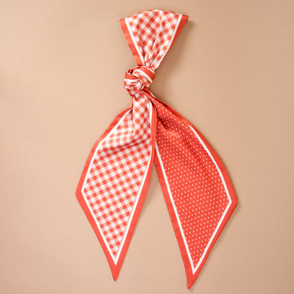 The Gingham Orange Twirl & Tie Scarf Accessory by The Darling Effect is displayed on a neutral beige background, featuring a vibrant orange and white satin design with gingham pattern on one side and polka dots on the other—perfect as a versatile accessory to elevate your styling options.