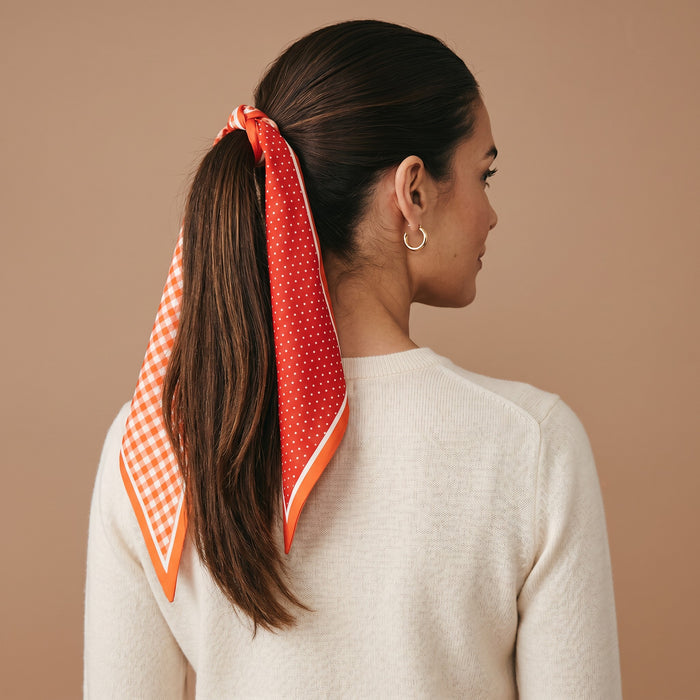 A woman with long brown hair styled in a ponytail, tied with The Darling Effect’s Gingham Orange Twirl & Tie Scarf Accessory featuring polka dot and gingham patterns. She wears a cream sweater and gold hoop earrings, facing away from the camera against a beige background.