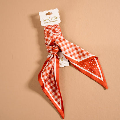 The Gingham Orange Twirl & Tie Scarf Accessory by The Darling Effect showcases a vibrant orange and white satin design with playful checkered and polka dot patterns. Elegantly presented on a branded cardboard holder against a beige background, this versatile accessory is perfect for enhancing your wardrobe with multiple styling options.