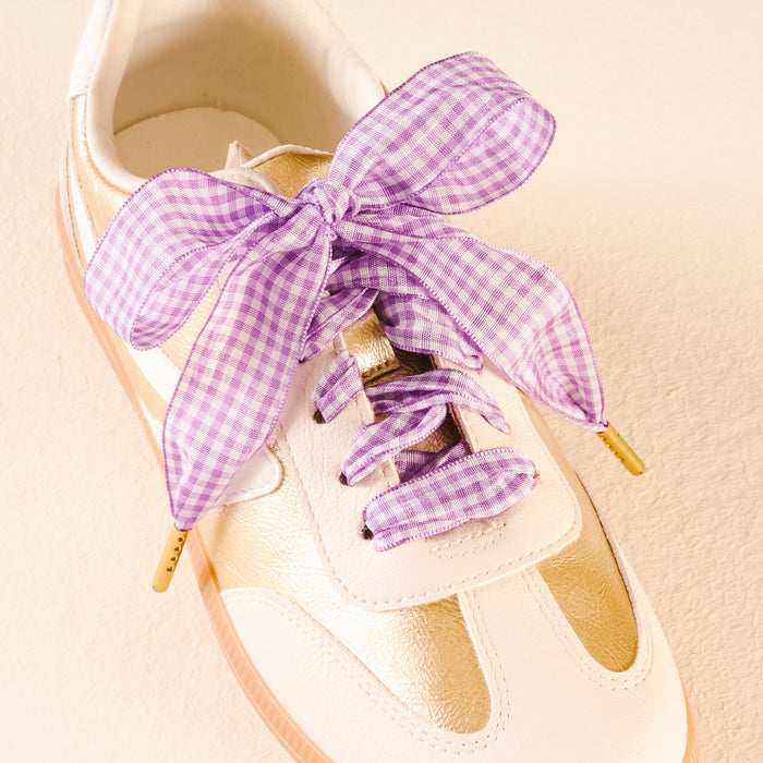 The Darling Effect's Gingham Purple Step It Up Shoelaces, wide with a purple gingham pattern and tied in a large bow, are styled on a white and gold sneaker against a light beige background.