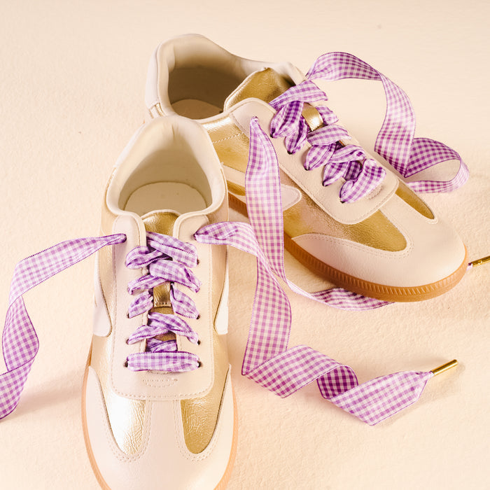 Two cream and gold sneakers with gum soles are shown on a light surface, featuring Gingham Purple Step It Up Shoelaces by The Darling Effect—wide, purple and white checkered laces loosely tied for a stylish touch.