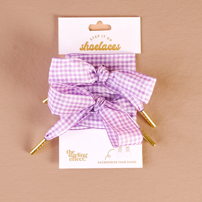 A pair of Gingham Purple Step It Up Shoelaces by The Darling Effect, featuring lilac and white checks with gold aglets, tied in bows on a beige background. Packaging says "Step It Up shoelaces" and "Accessorize your shoes.