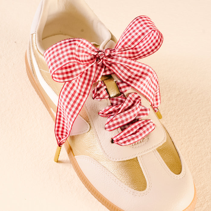 A white and gold sneaker is laced with The Darling Effect’s Gingham Red Step It Up Shoelaces, featuring a red and white gingham ribbon tied in a large bow, displayed on a light beige surface.