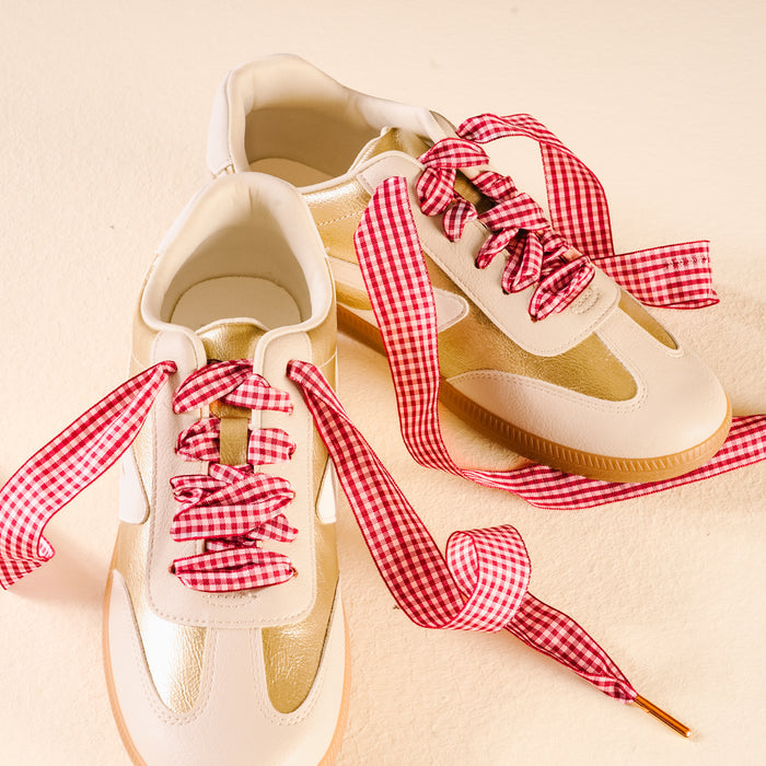A pair of white sneakers with gold accents, laced with The Darling Effect's Gingham Red Step It Up Shoelaces, displayed on a light cream-colored surface.