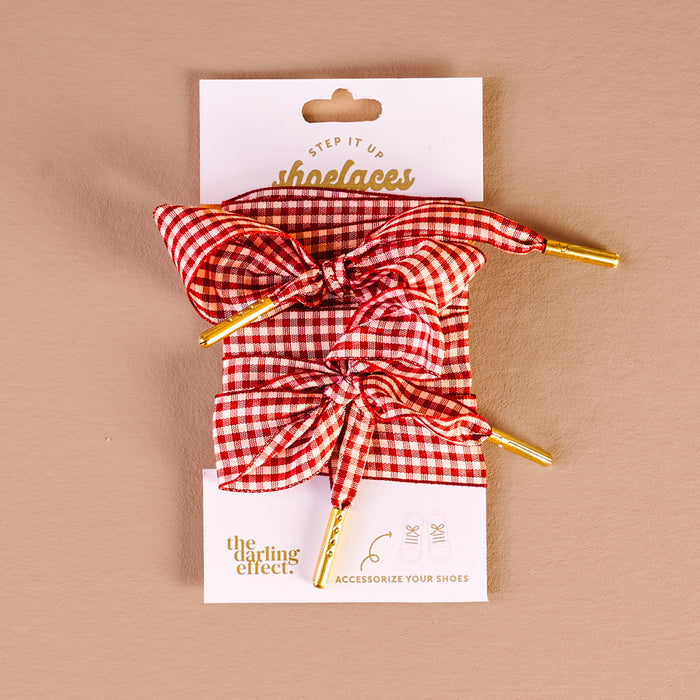Add a pop of style to your shoes with the Gingham Red Step It Up Shoelaces from The Darling Effect—red and white gingham laces with gold aglets, tied in bows and displayed on a branded card against a beige background.