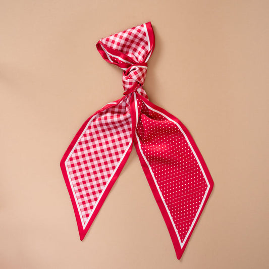 The Darling Effect's Gingham Red Twirl & Tie Scarf Accessory showcases a vibrant red and white checkered pattern on one side and playful polka dots on the other, styled in a loose knot against a beige background—ideal for experimenting with versatile looks.