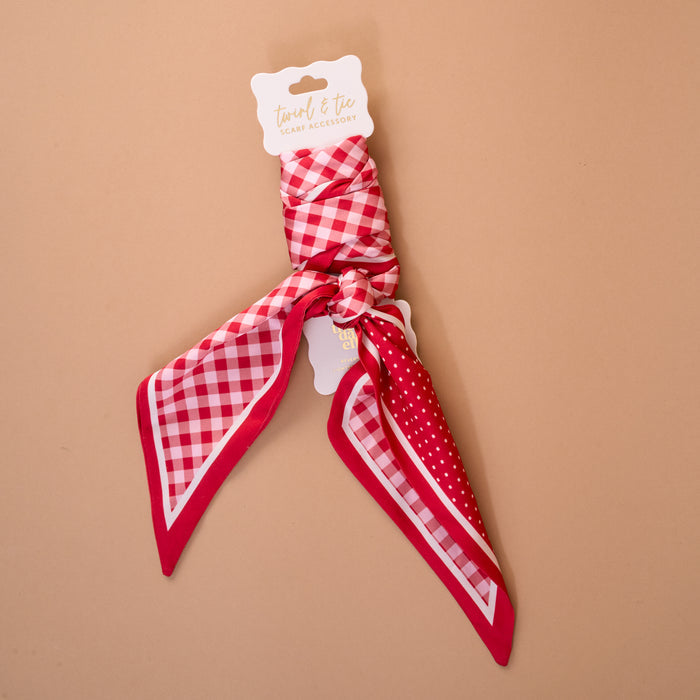 The Gingham Red Twirl & Tie Scarf Accessory by The Darling Effect features red satin adorned with white checkered and polka dot patterns, elegantly tied around a white card on a light brown background, providing versatile styling options for any look.