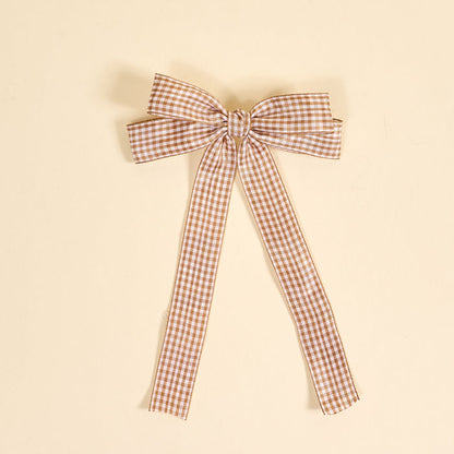 The Darling Effect's Gingham Tan Double Take Hair Bow features a brown and white checkered ribbon with long tails, perfect as a stylish hair accessory. Displayed on a plain beige background.