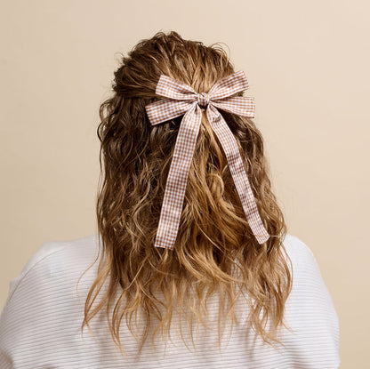 A person with wavy, shoulder-length hair is shown from behind in a white textured top. Their hair is styled half-up and secured with The Darling Effect’s Gingham Tan Double Take Hair Bow—a charming beige-pink accessory.