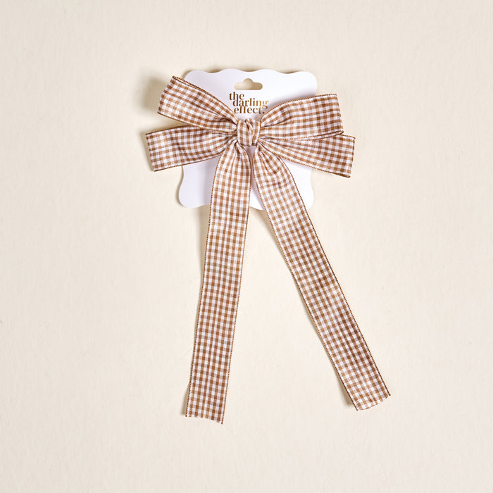 The Gingham Tan Double Take Hair Bow by The Darling Effect features a brown and white gingham pattern with long tails, displayed on a branded card—an adorable addition to your hair accessory collection.