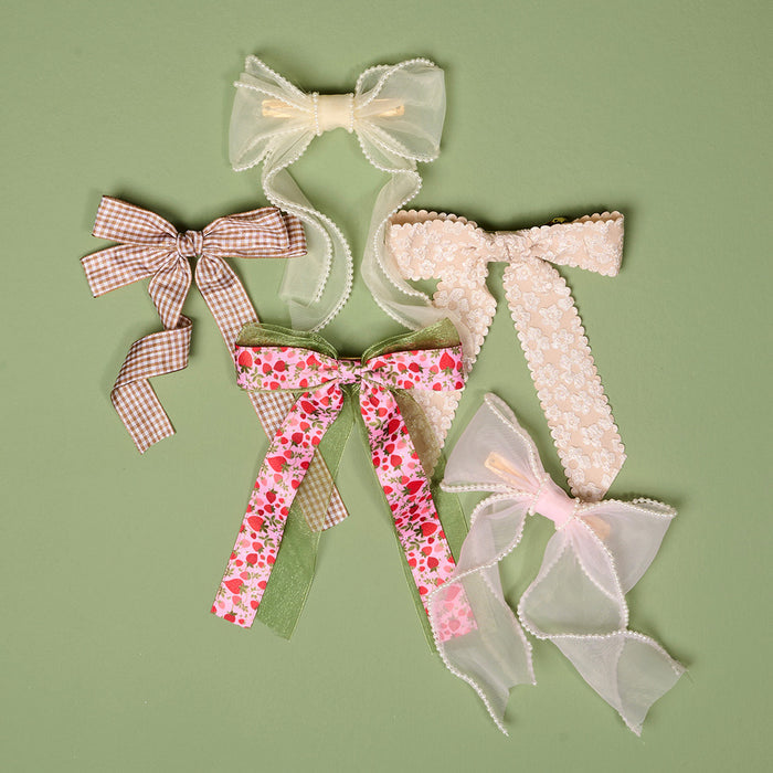 The Darling Effect’s Gingham Tan Double Take Hair Bow is a charming hair accessory, beautifully displayed among other bows made of lace, sheer white, and pink floral fabrics on a light green background.