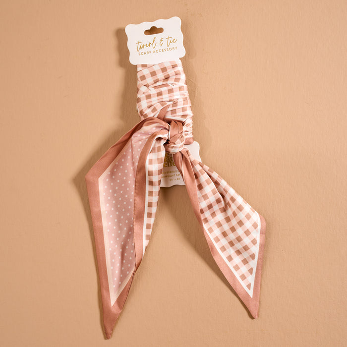 The Darling Effect’s Gingham Tan Twirl & Tie Scarf Accessory is a pink and white scrunchie with an attached satin scarf, featuring checkered and polka dot patterns, displayed on a tan background with a “twirl & tie” label—perfect for creating multiple chic styling options.