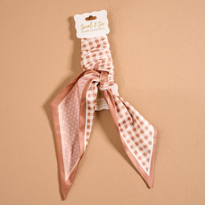 The Darling Effect’s Gingham Tan Twirl & Tie Scarf Accessory is a pink and white scrunchie with an attached satin scarf, featuring checkered and polka dot patterns, displayed on a tan background with a “twirl & tie” label—perfect for creating multiple chic styling options.