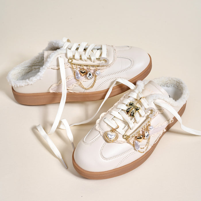 A pair of white slip-on sneakers with faux fur lining and tan soles are styled with The Darling Effect’s Glitz & Glam Dazzle Shoe Charm Pack—gold and pearl heart and bow charms—making them playful accessories, shown on a light background.
