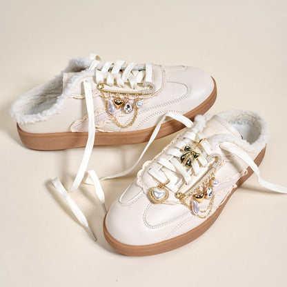 A pair of white slip-on sneakers with faux fur lining and tan soles are styled with The Darling Effect’s Glitz & Glam Dazzle Shoe Charm Pack—gold and pearl heart and bow charms—making them playful accessories, shown on a light background.