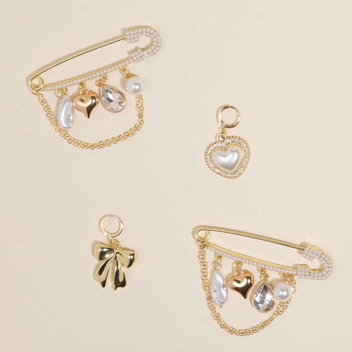 The Darling Effect’s Glitz & Glam Dazzle Shoe Charm Pack features three gold safety pins with pearl, heart, and crystal charms, plus two separate charms—a pearl heart and gold bow—arranged elegantly on a light beige background.