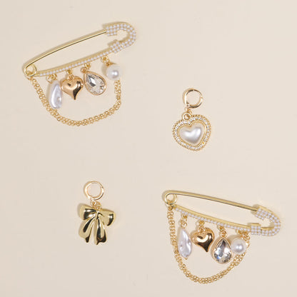 The Darling Effect’s Glitz & Glam Dazzle Shoe Charm Pack features three gold safety pins with pearl, heart, and crystal charms, plus two separate charms—a pearl heart and gold bow—arranged elegantly on a light beige background.