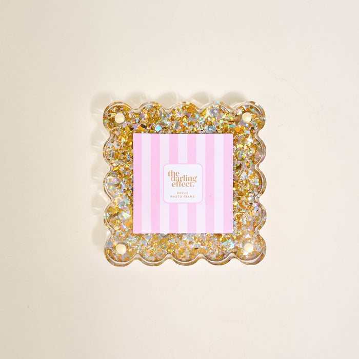 The Gold Glitter Little Moments Acrylic Picture Frame by The Darling Effect features a square, scalloped-edge design with gold and silver glitter accents, shown with a pink and white striped card on a light background.