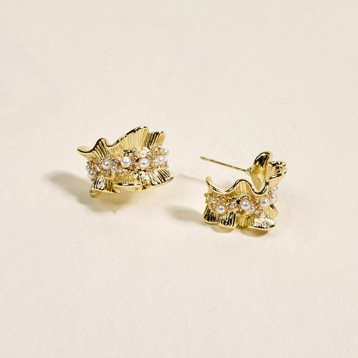 The Darling Effect’s Golden Ruffle Standout Earrings shine with a delicate leaf design, accented by white pearls and crystals, and are shown against a light beige background.