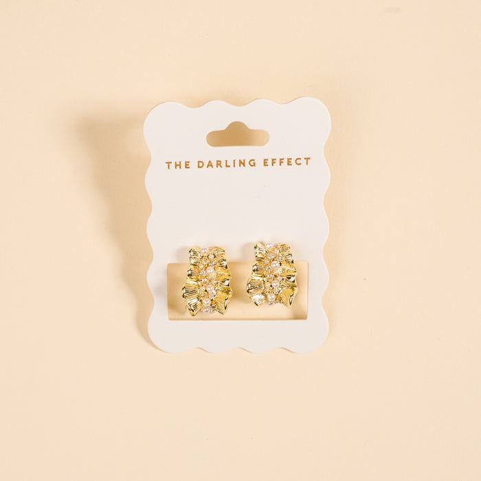 Golden Ruffle Standout Earrings by The Darling Effect feature textured gold studs with pearl and crystal accents, displayed on a white scalloped card against a soft peach background.