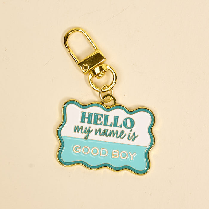 The Darling Effect’s Good Boy Pawfect Collar Charm features a gold keychain with a sign-shaped charm that reads "HELLO my name is GOOD BOY" in white, teal, and gold—a playful way to showcase your pet’s unique style.