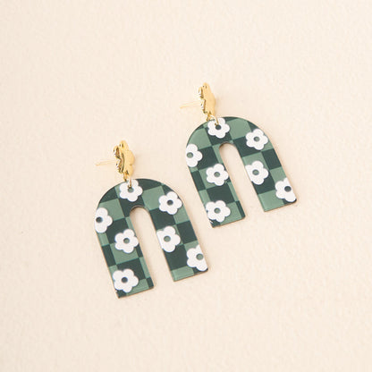 Show off your school colors with The Darling Effect's Green Game Day Acrylic Earrings—arch-shaped, featuring a green and black checkered pattern, white floral accents, and gold-tone studs—photographed on a light background.