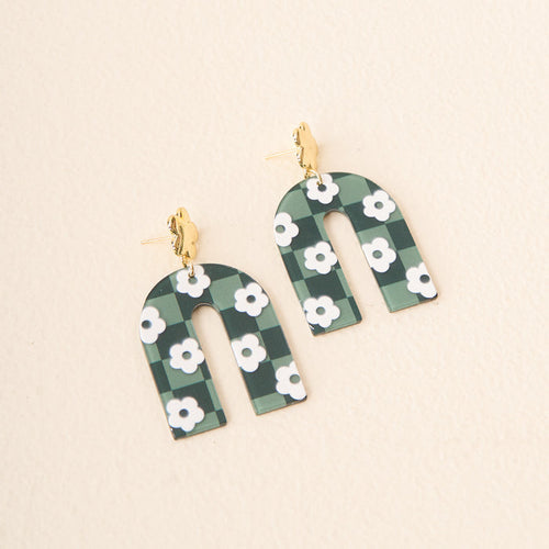 Show off your school colors with The Darling Effect's Green Game Day Acrylic Earrings—arch-shaped, featuring a green and black checkered pattern, white floral accents, and gold-tone studs—photographed on a light background.