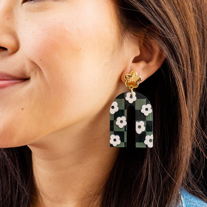 A woman smiles wearing The Darling Effect's Green Game Day Acrylic Earrings—arch-shaped with a green and black checkered pattern, white flowers, and gold flower studs—perfect for showing school spirit.