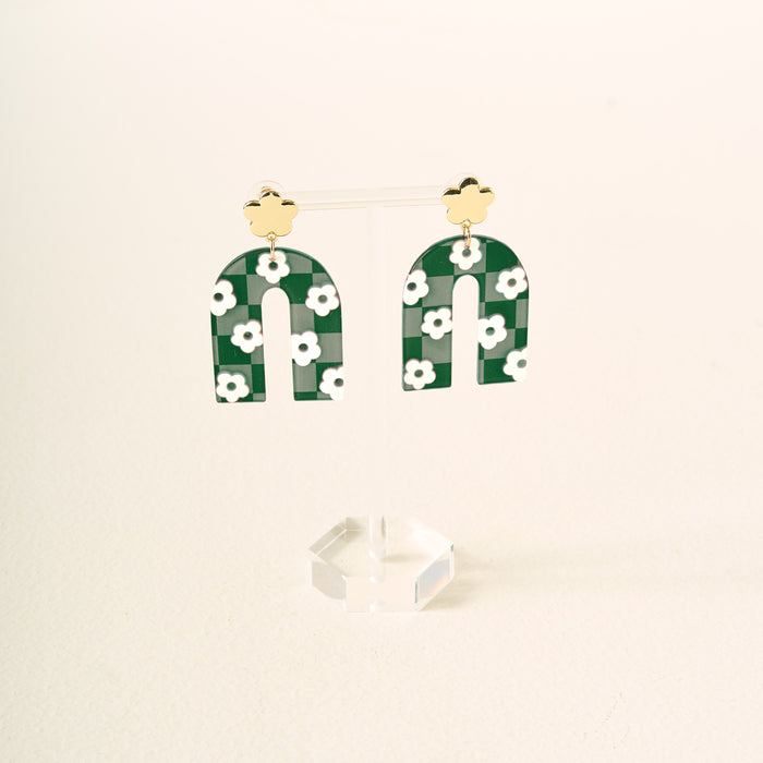 Show off your school colors with the Green Game Day Acrylic Earrings by The Darling Effect—green arch-shaped acrylic earrings with white flowers, hanging from gold flower studs and displayed on a clear stand against a light background.