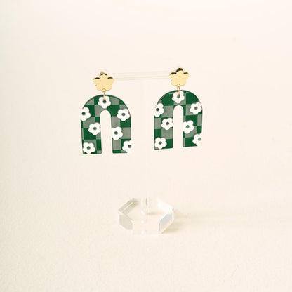 Show off your school colors with the Green Game Day Acrylic Earrings by The Darling Effect—green arch-shaped acrylic earrings with white flowers, hanging from gold flower studs and displayed on a clear stand against a light background.