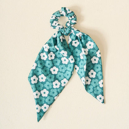 The Darling Effect’s Green Game Day Hair Scarf with Scrunchie, featuring two ribbon tails and a daisy floral pattern, is displayed on an off-white background—ideal for showcasing your school colors or team spirit on game day.