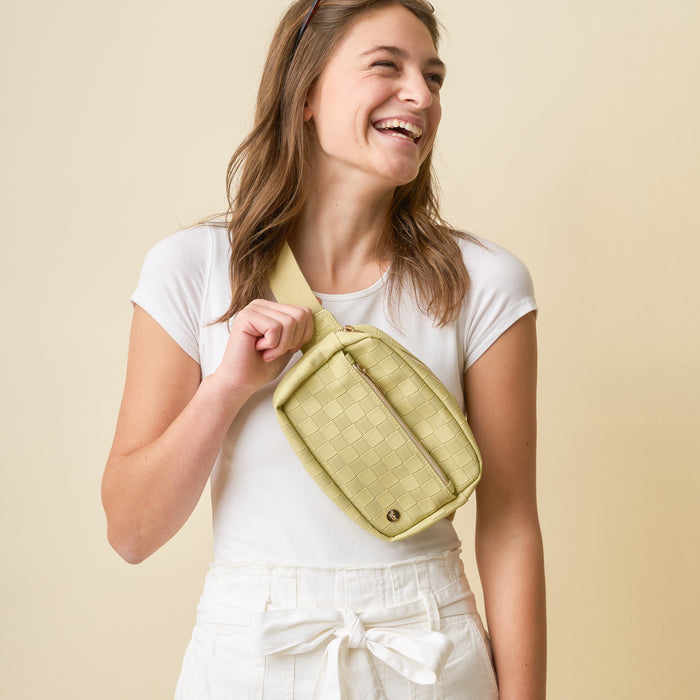 A smiling woman with long brown hair wears a white t-shirt and high-waisted white pants, posing with The Darling Effect's Green Urban Check Belt Bag against a beige background.
