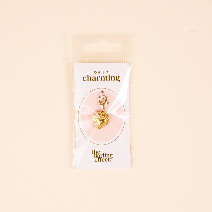 The Heart Jewelry Charm Gold by the-darling-effect features a gold heart-shaped charm with a clasp, displayed on a white Oh So Charming card and presented in clear packaging against a beige background.