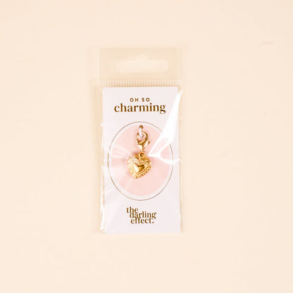 The Heart Jewelry Charm Gold by the-darling-effect features a gold heart-shaped charm with a clasp, displayed on a white Oh So Charming card and presented in clear packaging against a beige background.